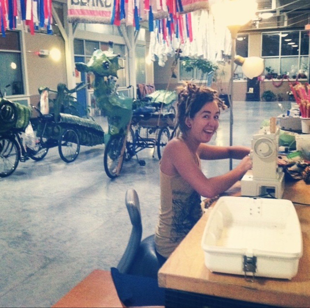 Young woman smiling at sewing machine in a bright, decorated indoor space with bicycles and colorful banners.
