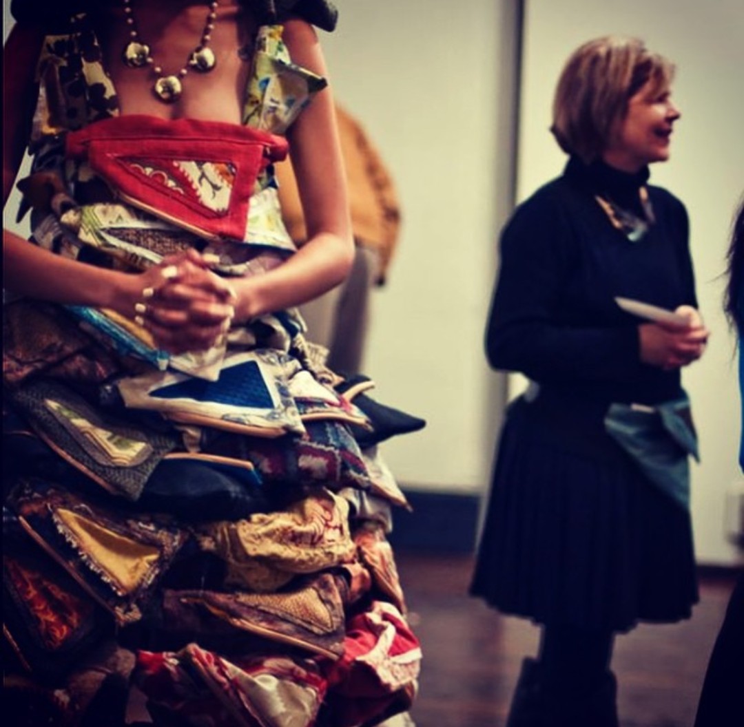 Person wearing a colorful, layered skirt made of fabric pouches, with hands clasped in front, and another person in black attire holding a book, in an indoor setting.