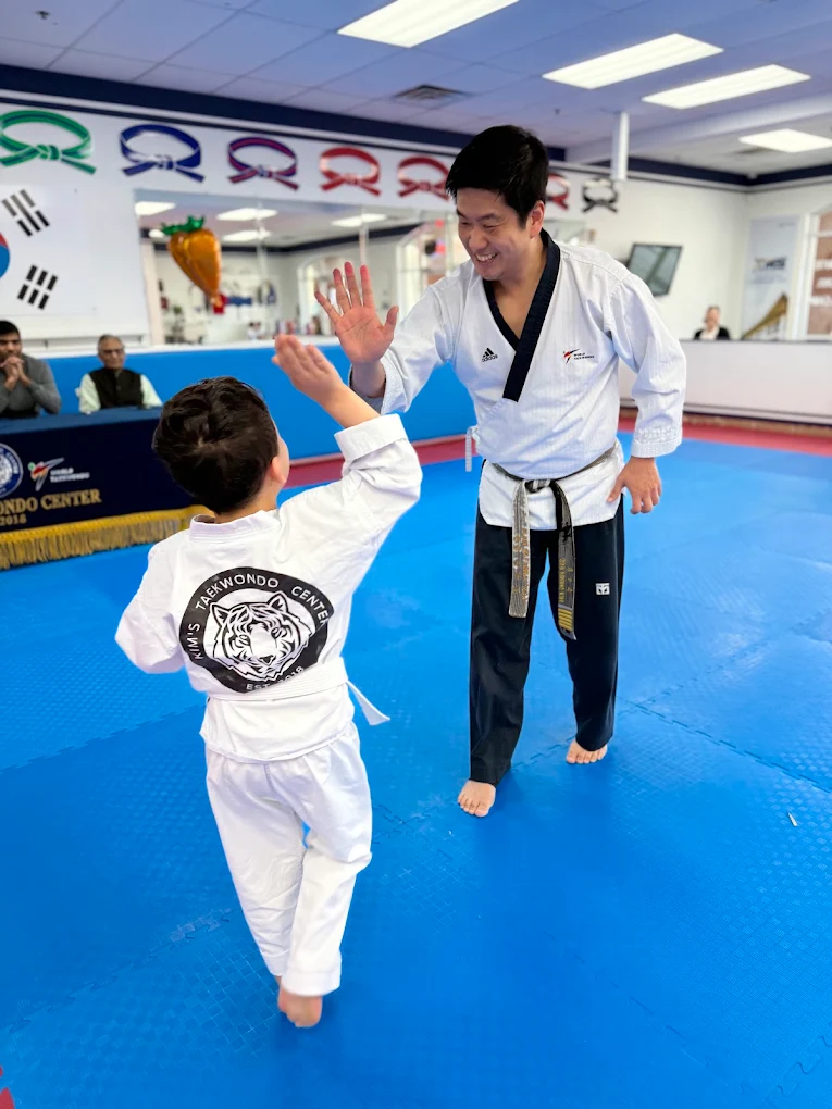 A martial arts instructor and a young student high-five in a dojo with blue mats and flags, smiling at each other.