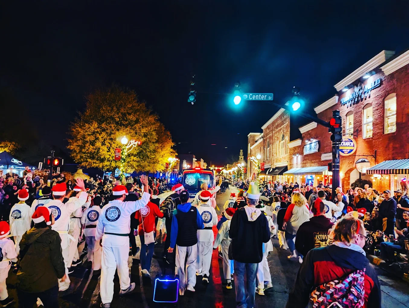 Crowd of people on a city street at night, some wearing Santa hats, with buildings and trees illuminated in the background.