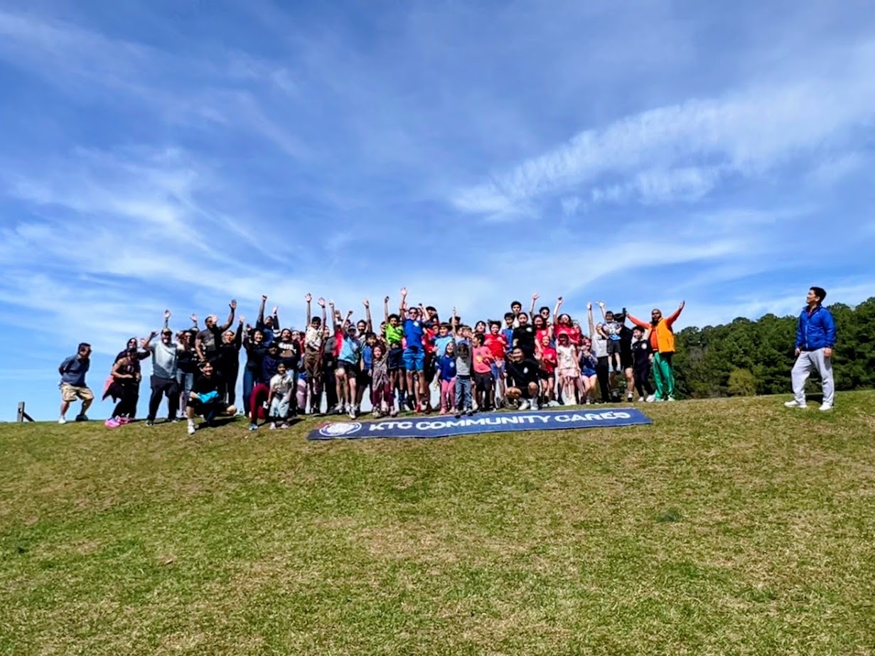 Group of people outdoors on a grassy hill under a blue sky, some raising their hands, with a banner on the ground.
