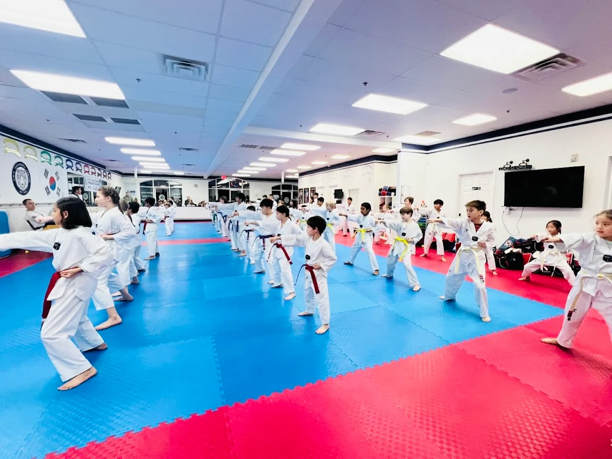 Children practicing martial arts in a brightly lit dojo with blue and red mats, wearing white uniforms with colored belts.