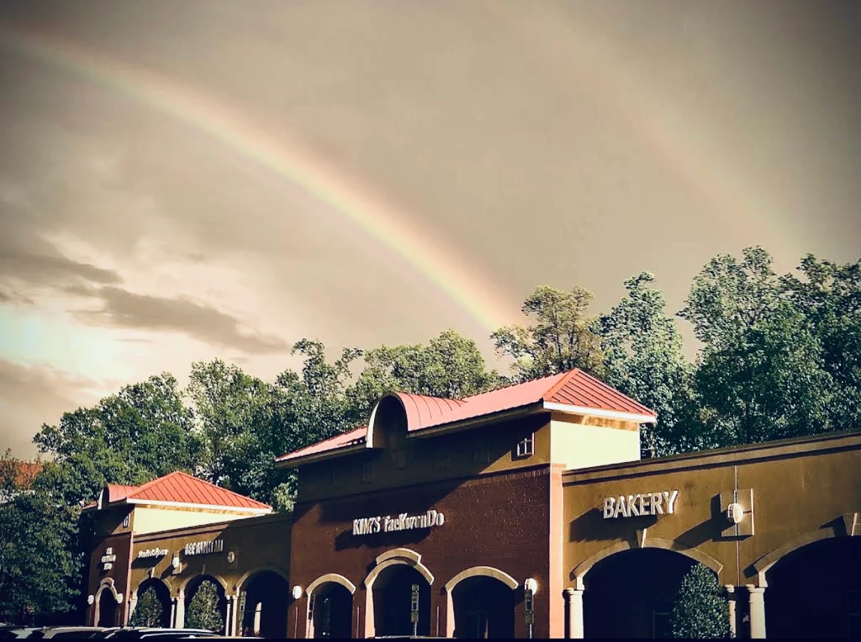 Rainbow arching over a bakery and storefront with trees in the background, sky partly cloudy, sunlight illuminating the scene.