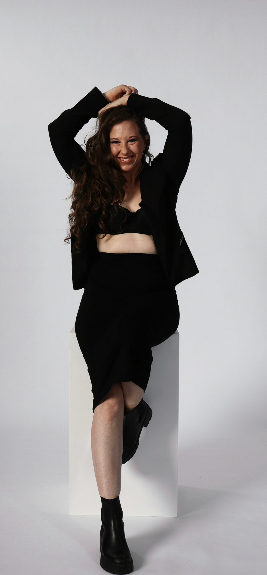 Woman with long curly hair smiling, wearing a black outfit and boots, standing with arms raised against a plain background.