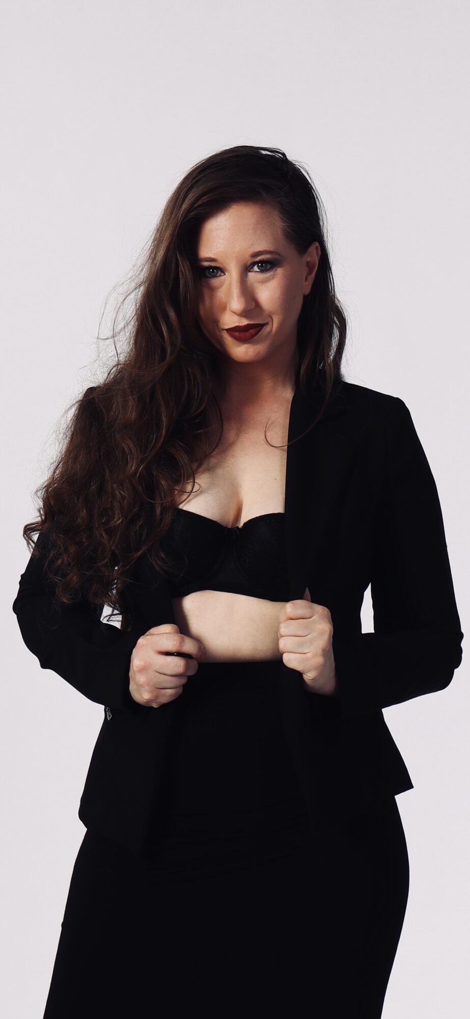 Woman with long dark hair wearing a black blazer and crop top, standing against a plain background.