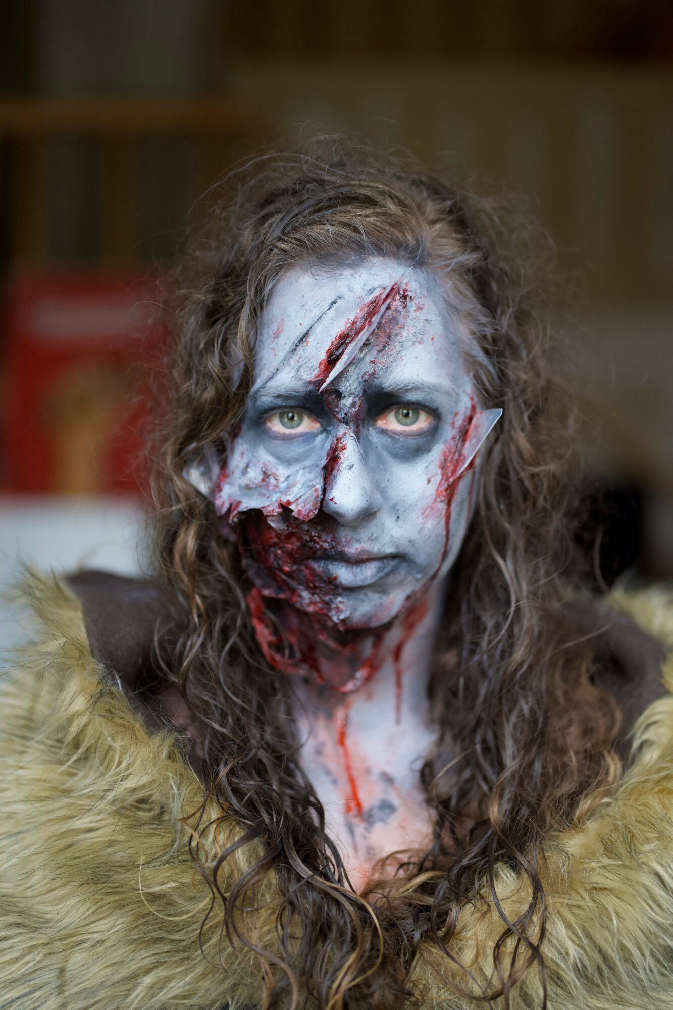 Person with face painted as a zombie, lying sideways, with makeup and contact lenses, in a room with blurred background.