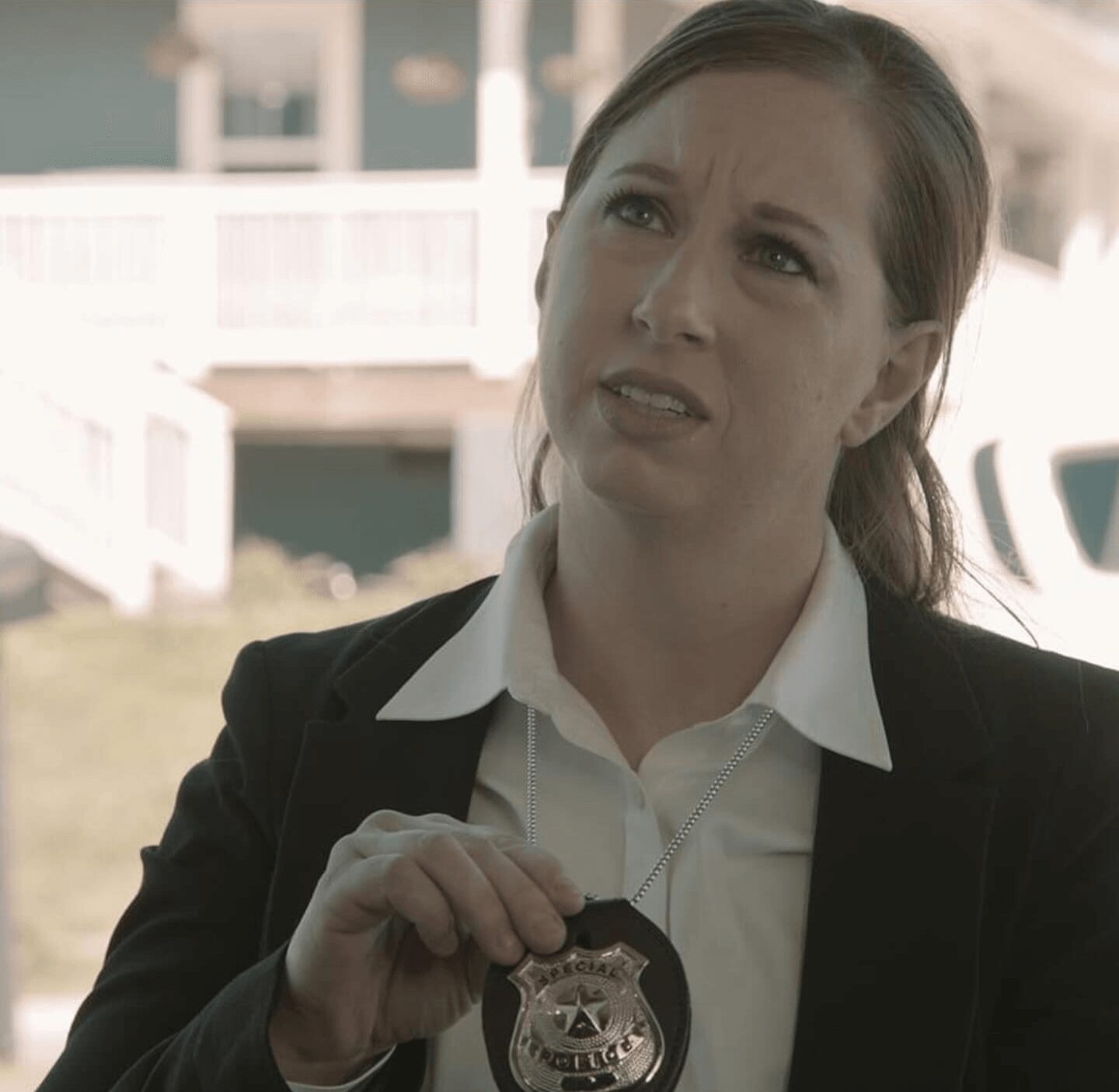 Woman in formal attire holding a police badge outdoors, with a house and porch in the background.