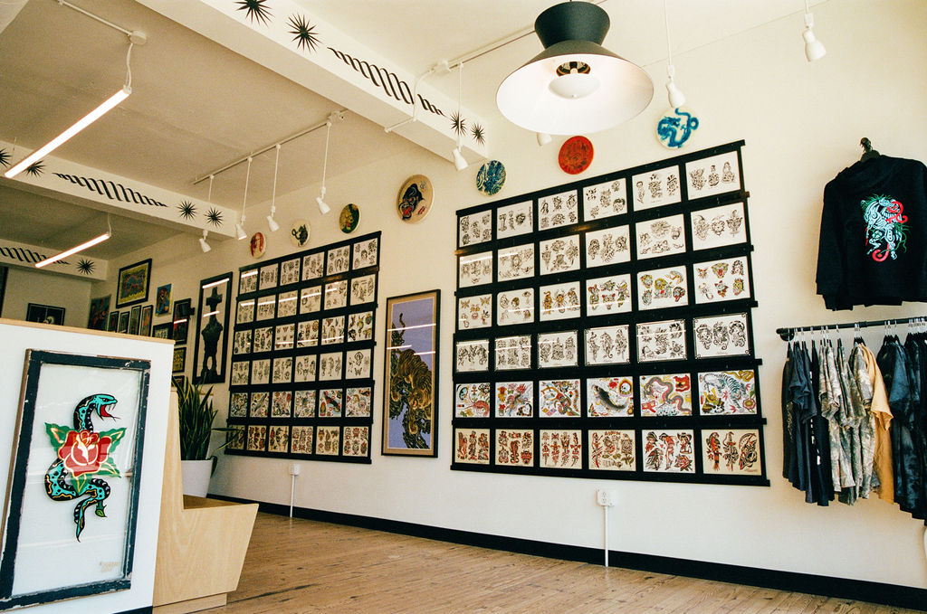 Art gallery with framed artwork on white walls, clothing display, and decorative plates on ceiling, with a hanging light fixture.