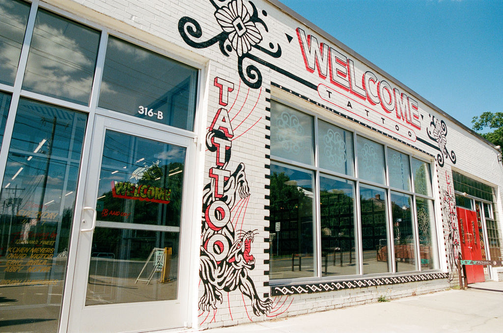 Tattoo shop storefront with large windows, decorative black and white floral designs, and a red and white welcome sign.