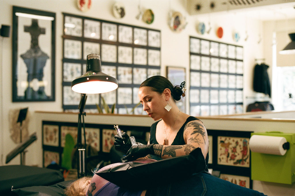 Tattooed woman with dark hair in bun, wearing black gloves, working on a client in a tattoo studio.