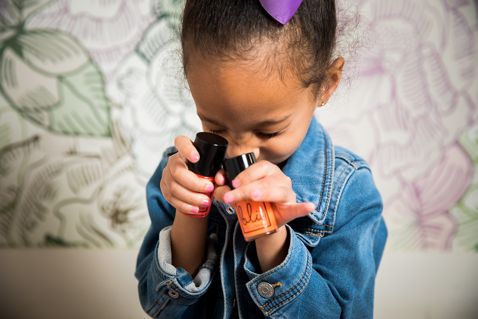 Young girl with a purple hair bow looking through a black and orange toy telescope, wearing a denim jacket, in front of a floral-patterned background.