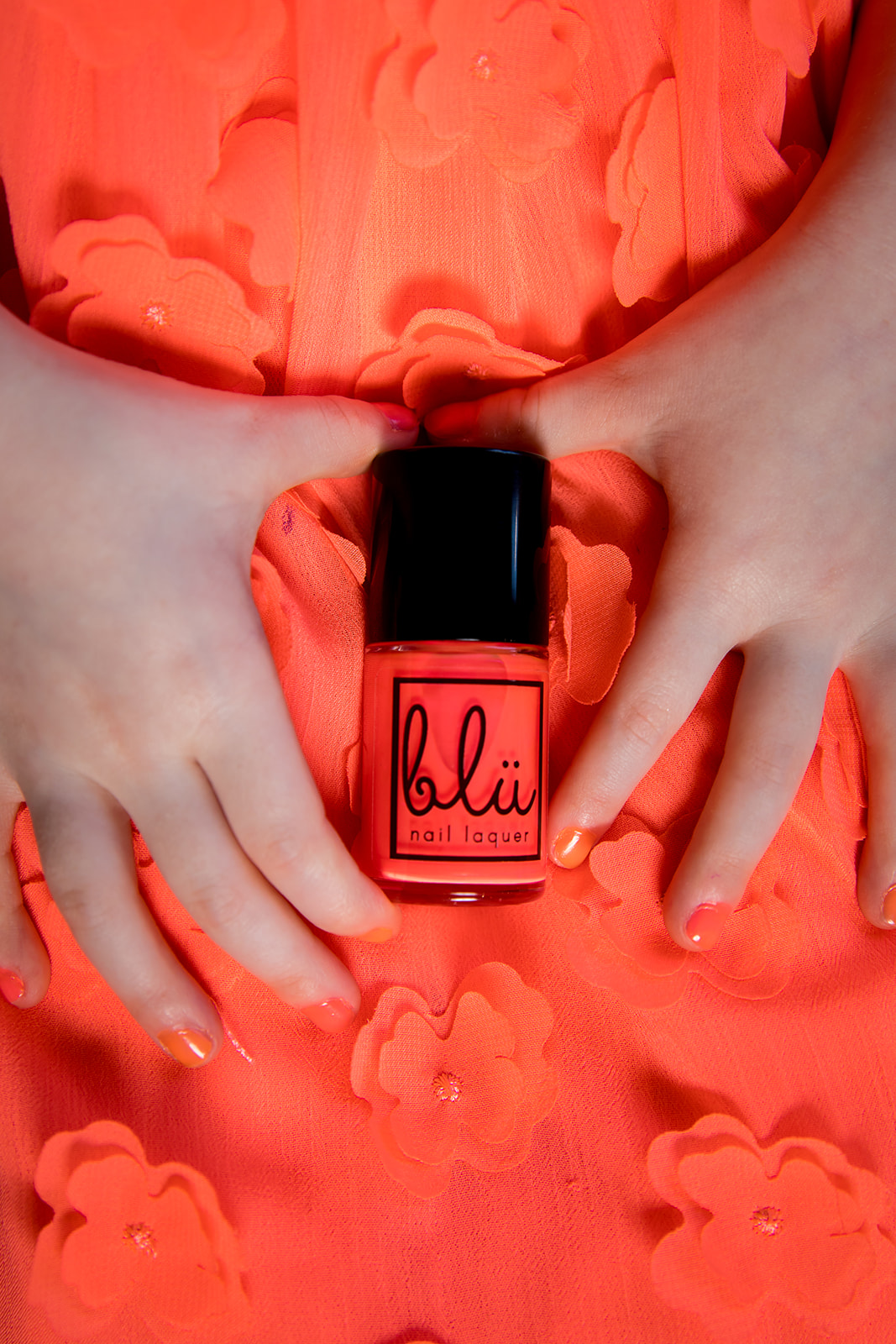 Two hands holding a coral-colored nail polish bottle on matching coral fabric background.