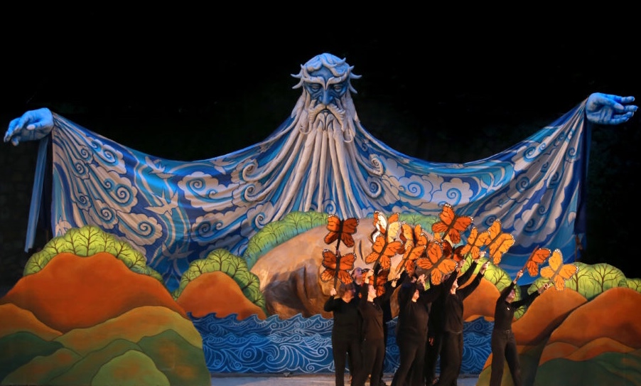 Stage scene with a large blue and white puppet resembling a mythical creature, surrounded by colorful scenery and performers.