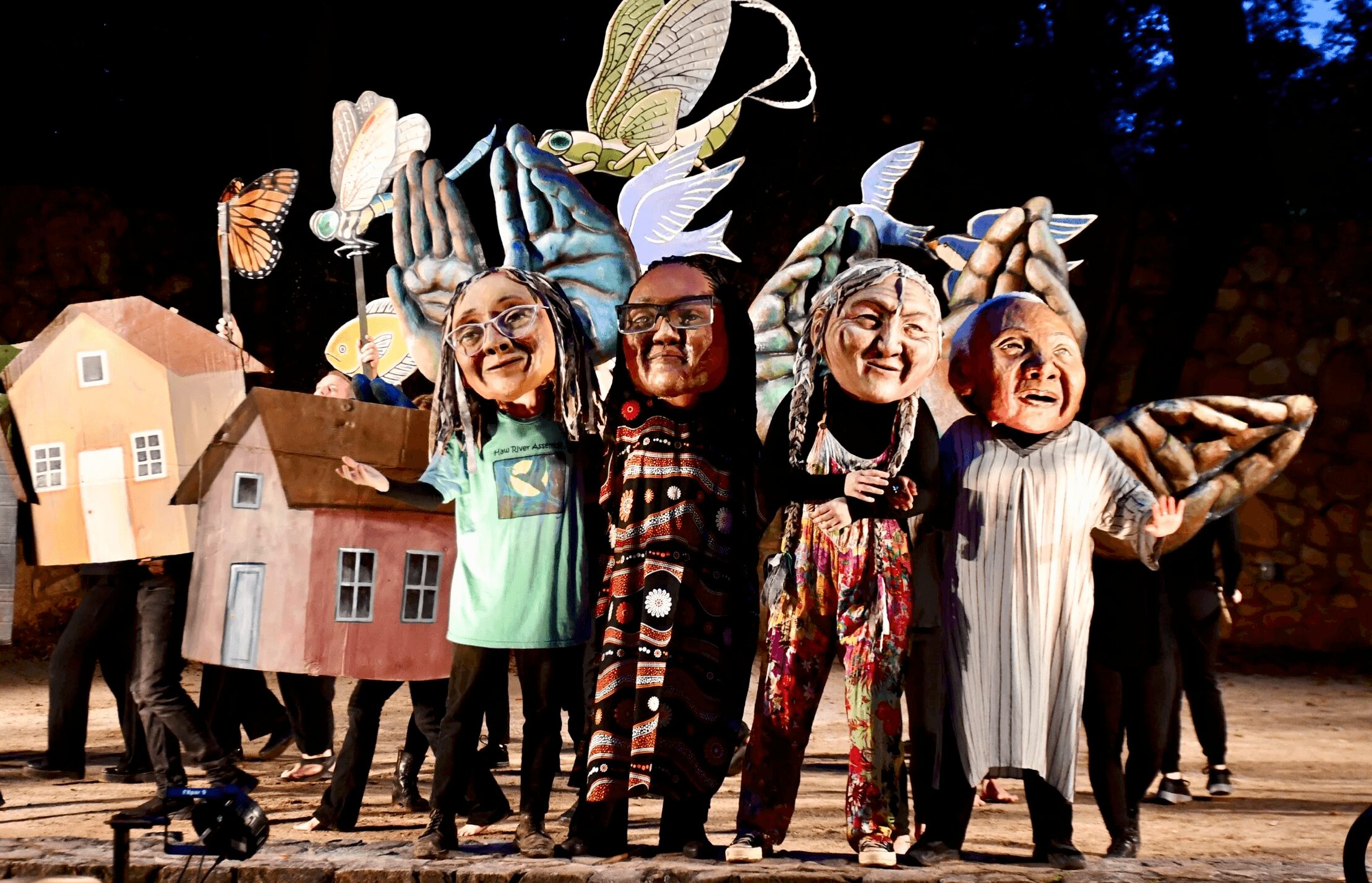 People in large puppet costumes perform on stage with trees in background, some holding houses and props, in a theatrical scene.