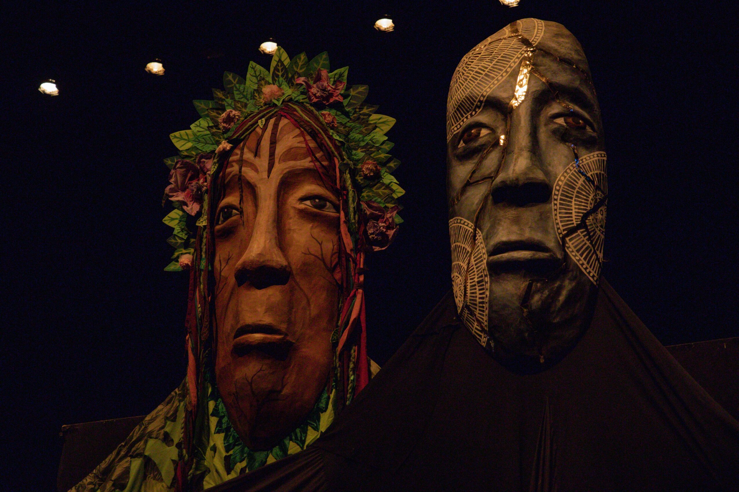 Two large masks or sculptures with expressive faces, one with a floral headdress, against a dark background.