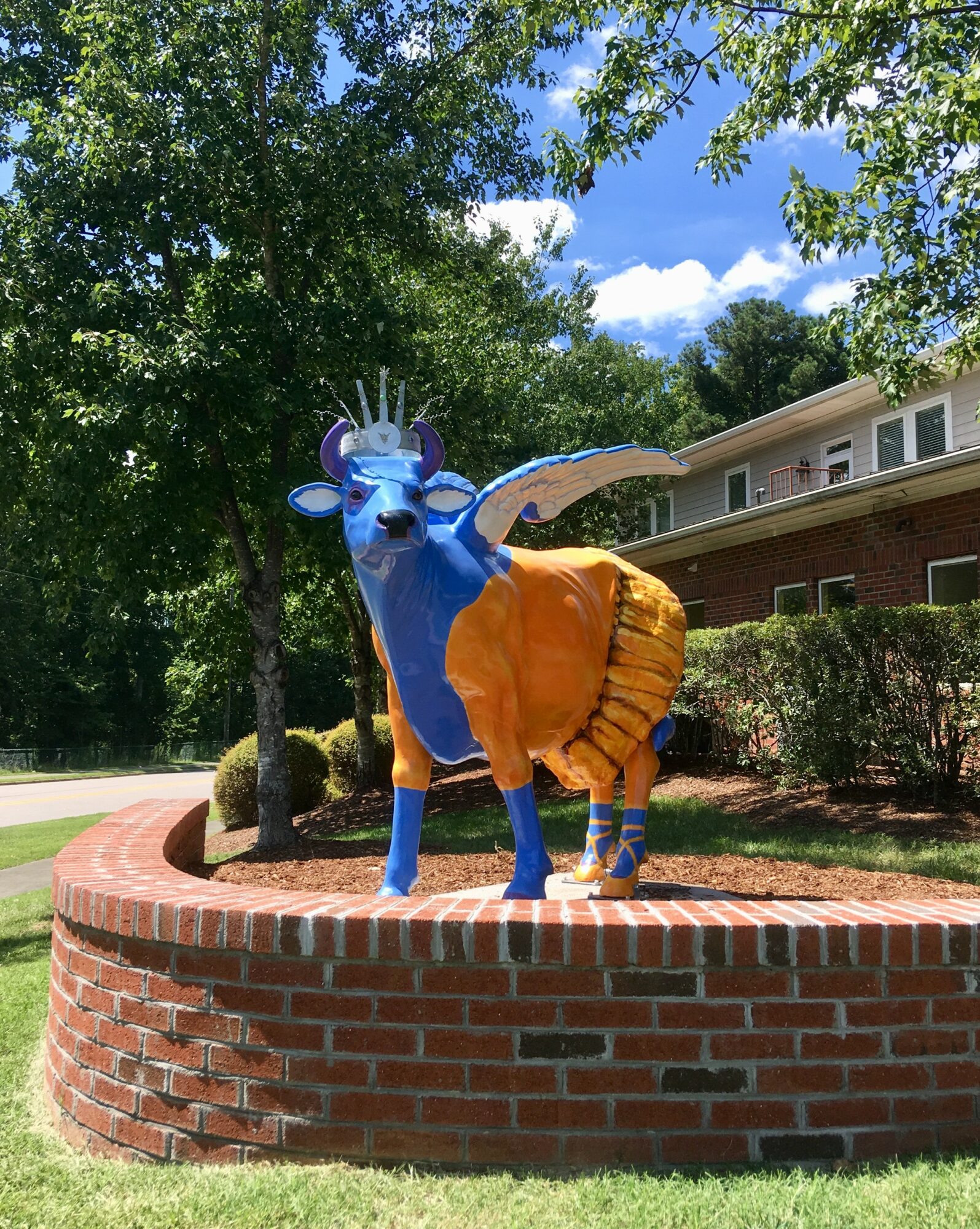 Colorful sculpture of a blue animal with orange clothing, standing on a brick circular platform outdoors, surrounded by trees and a building.
