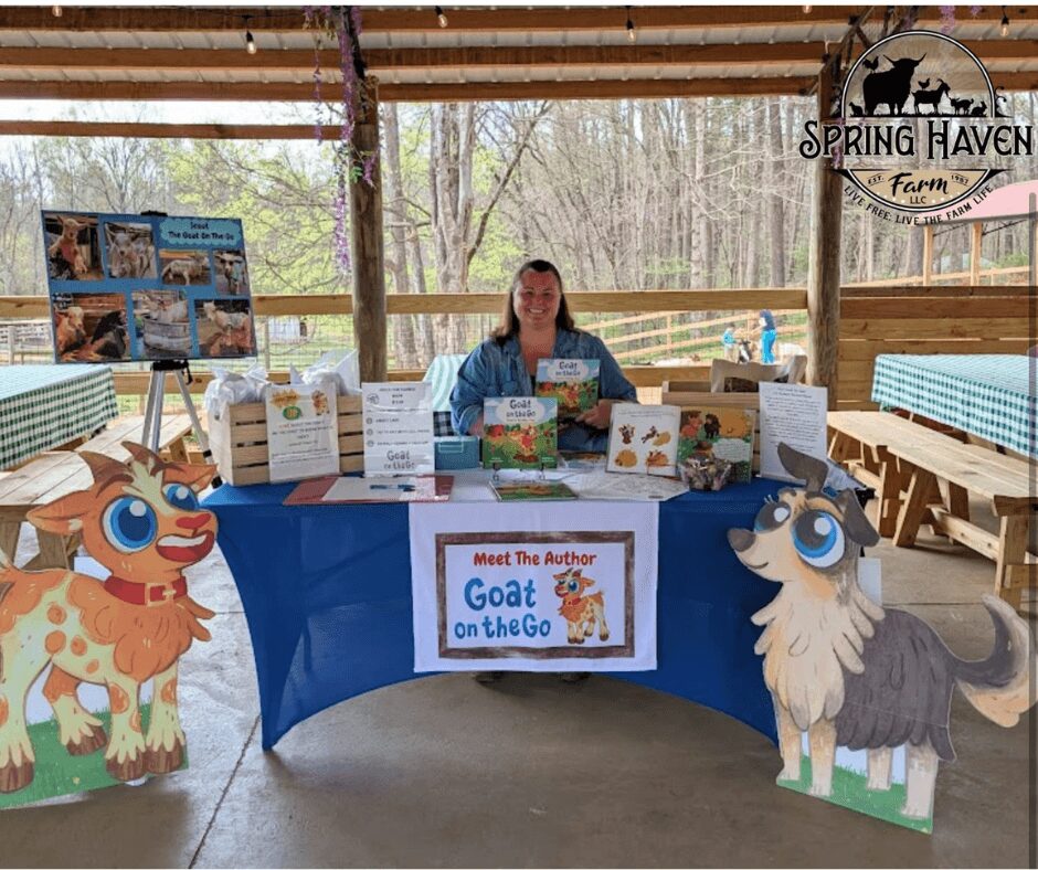 Woman sitting behind a table with books and signs, decorated with cartoon goat and sheep cutouts, at an outdoor farm event.
