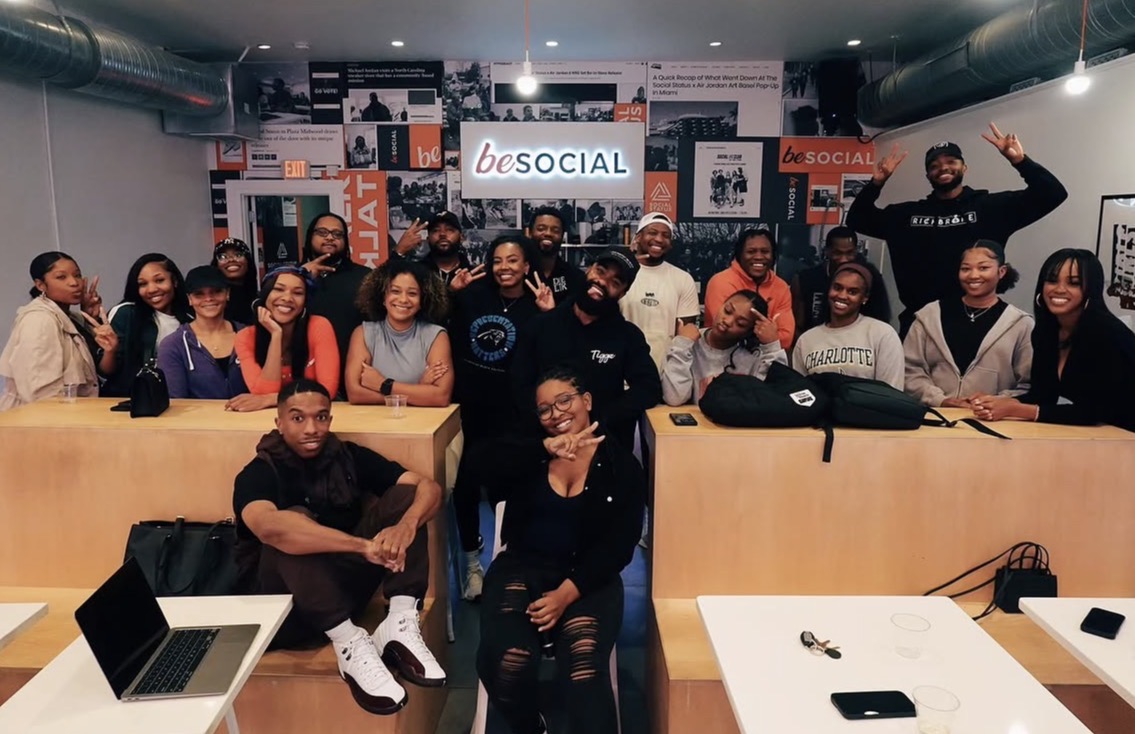 Group of people in a room with a screen displaying 'be SOCIAL' behind them, some sitting and some standing, smiling.
