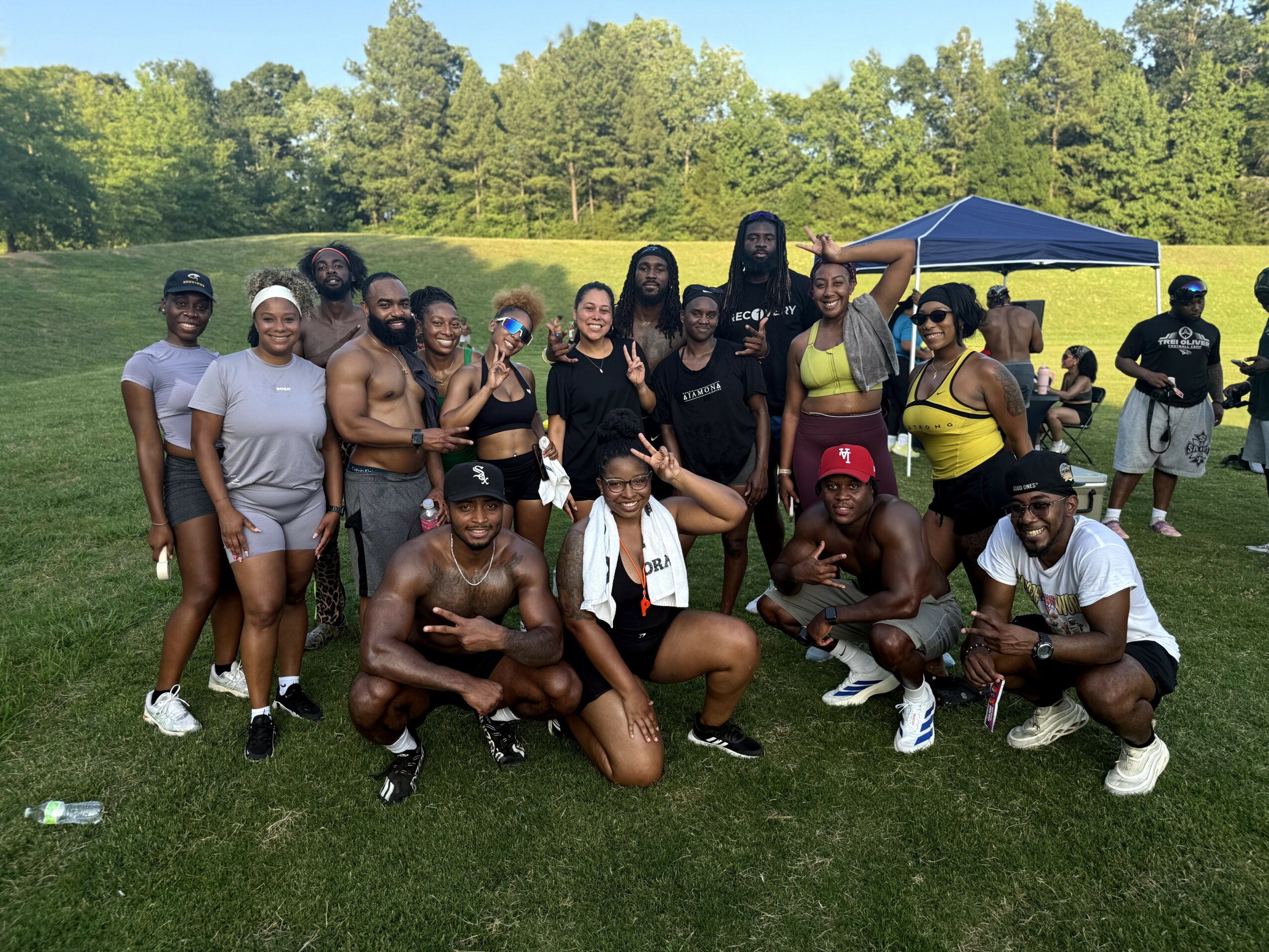 Group of people outdoors on grass, some shirtless, some wearing athletic clothing, posing for a photo with trees and a tent in background.
