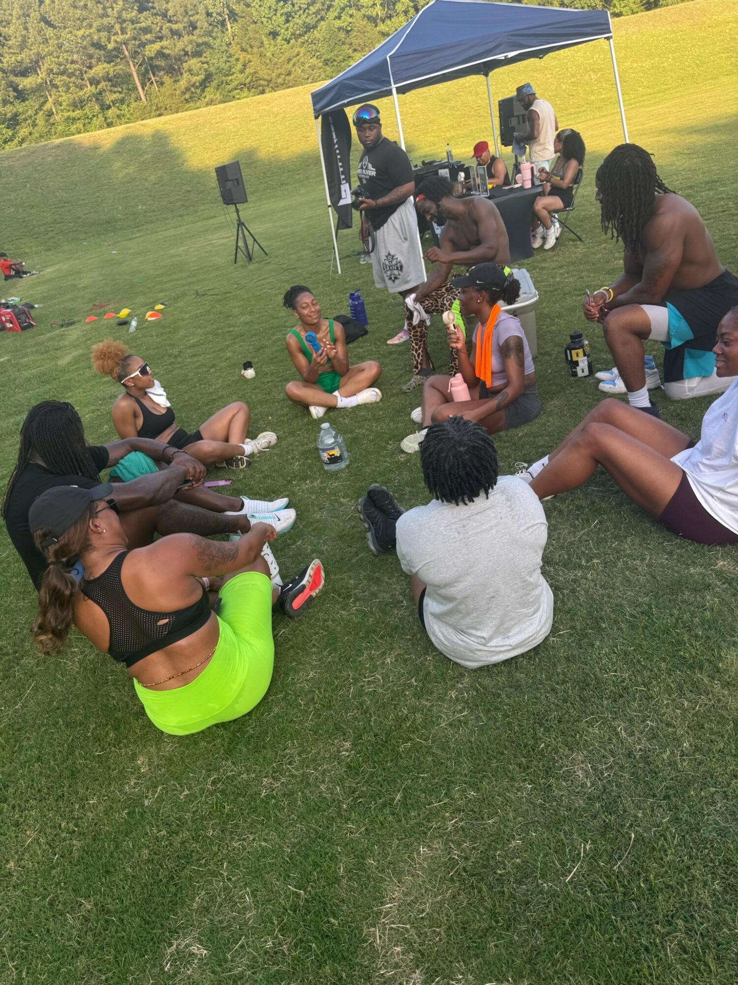 Group of people sitting and standing on grass during outdoor gathering with a canopy, drinks, and a speaker, in a park-like setting.
