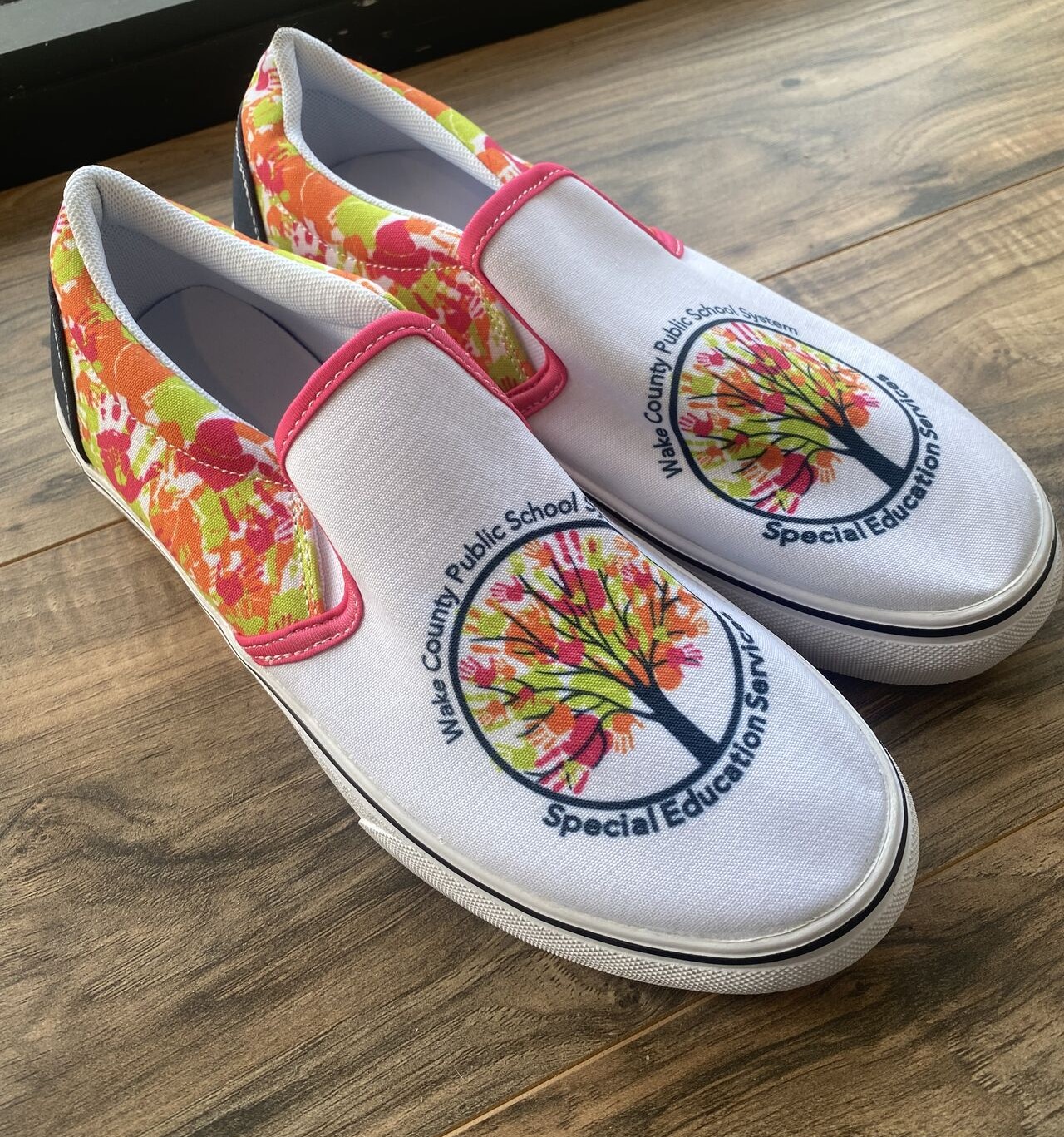 Pair of white slip-on shoes with colorful floral pattern on the sides and a logo of a tree with text around it.