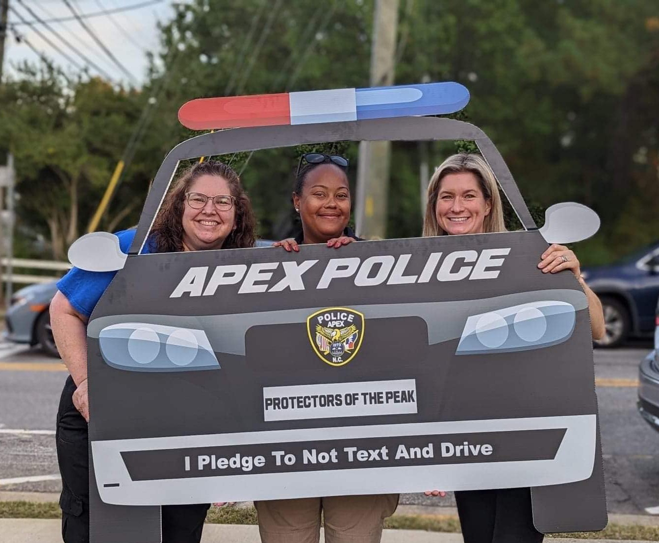 Three women holding a police-themed cardboard cutout with a police badge and text about not texting and driving.