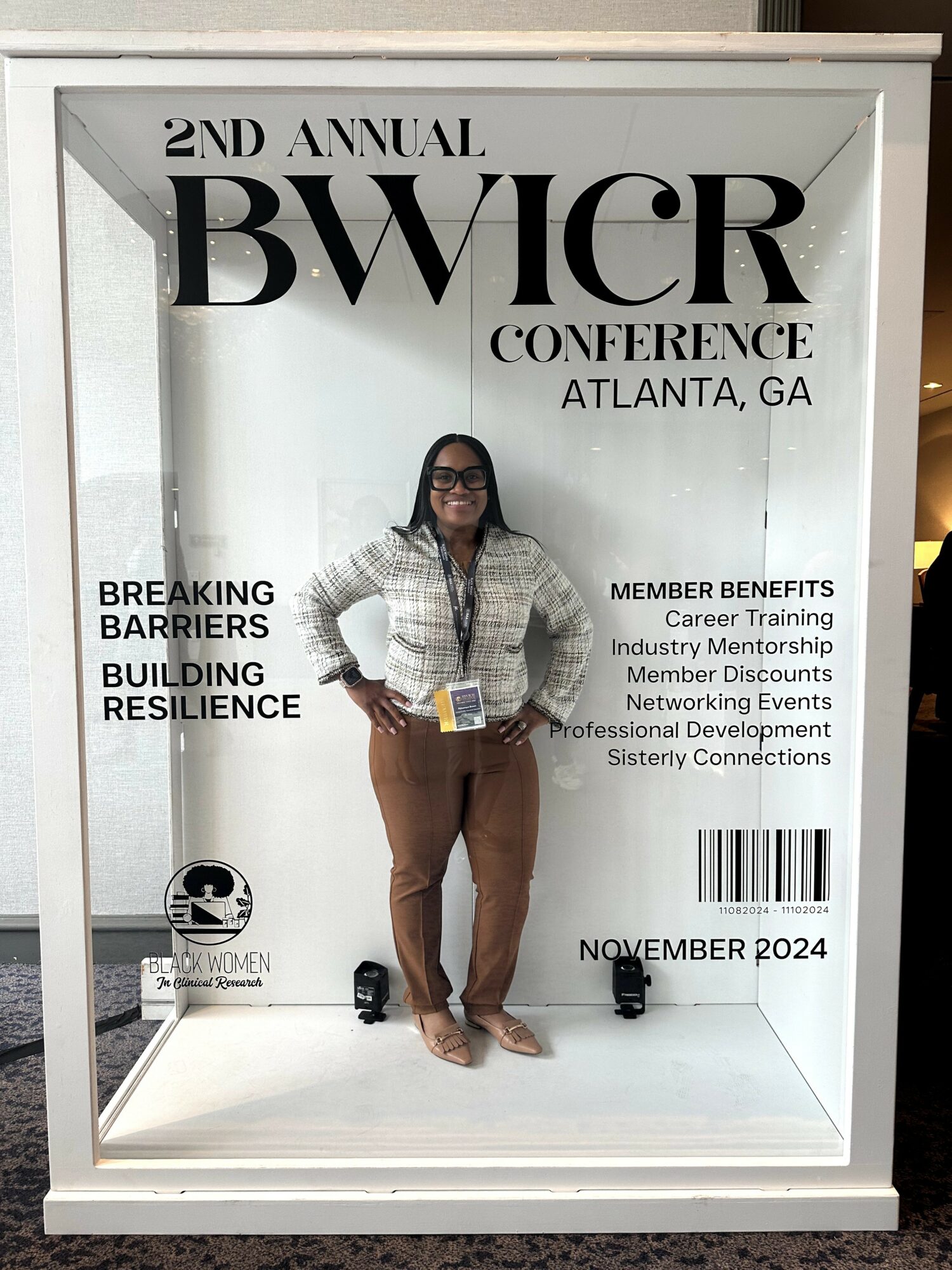 Woman standing in front of conference sign, holding a booklet, wearing glasses, a patterned top, and brown pants.