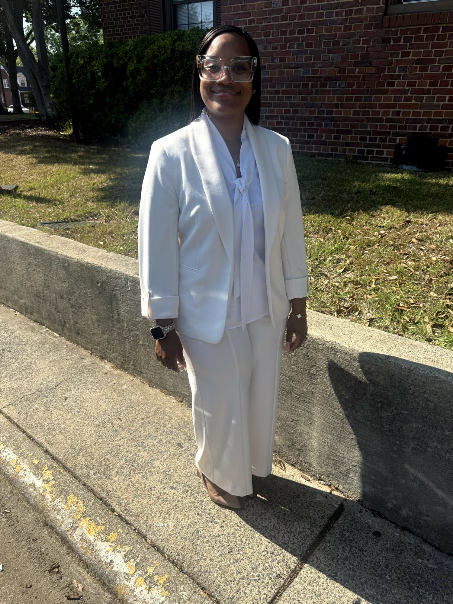 Woman in white suit standing outdoors on sidewalk, smiling, with brick building and greenery in background.