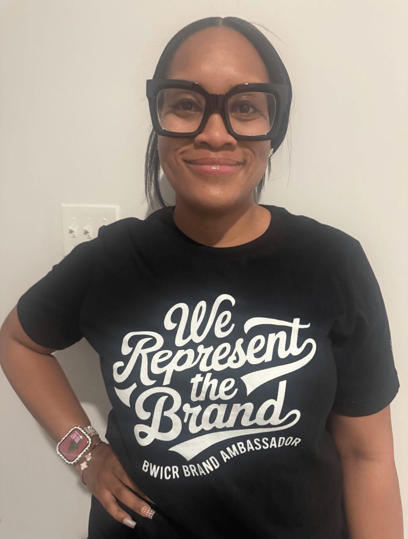 Woman wearing large glasses and a black T-shirt with white text, smiling, standing indoors against a plain wall.