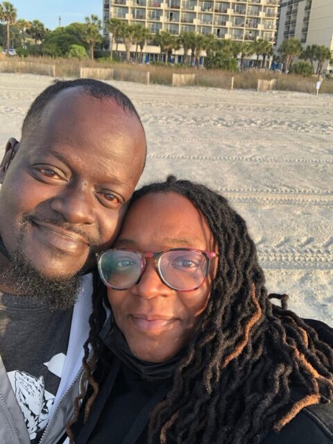 Two people smiling close together on a sandy beach with buildings in the background.