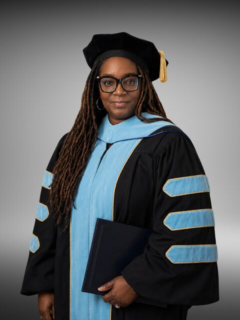 Woman in academic regalia holding a folder, wearing glasses and a graduation cap with a tassel, smiling.
