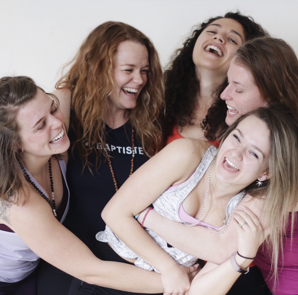 Five women laughing and hugging together, smiling joyfully, in a close group shot.