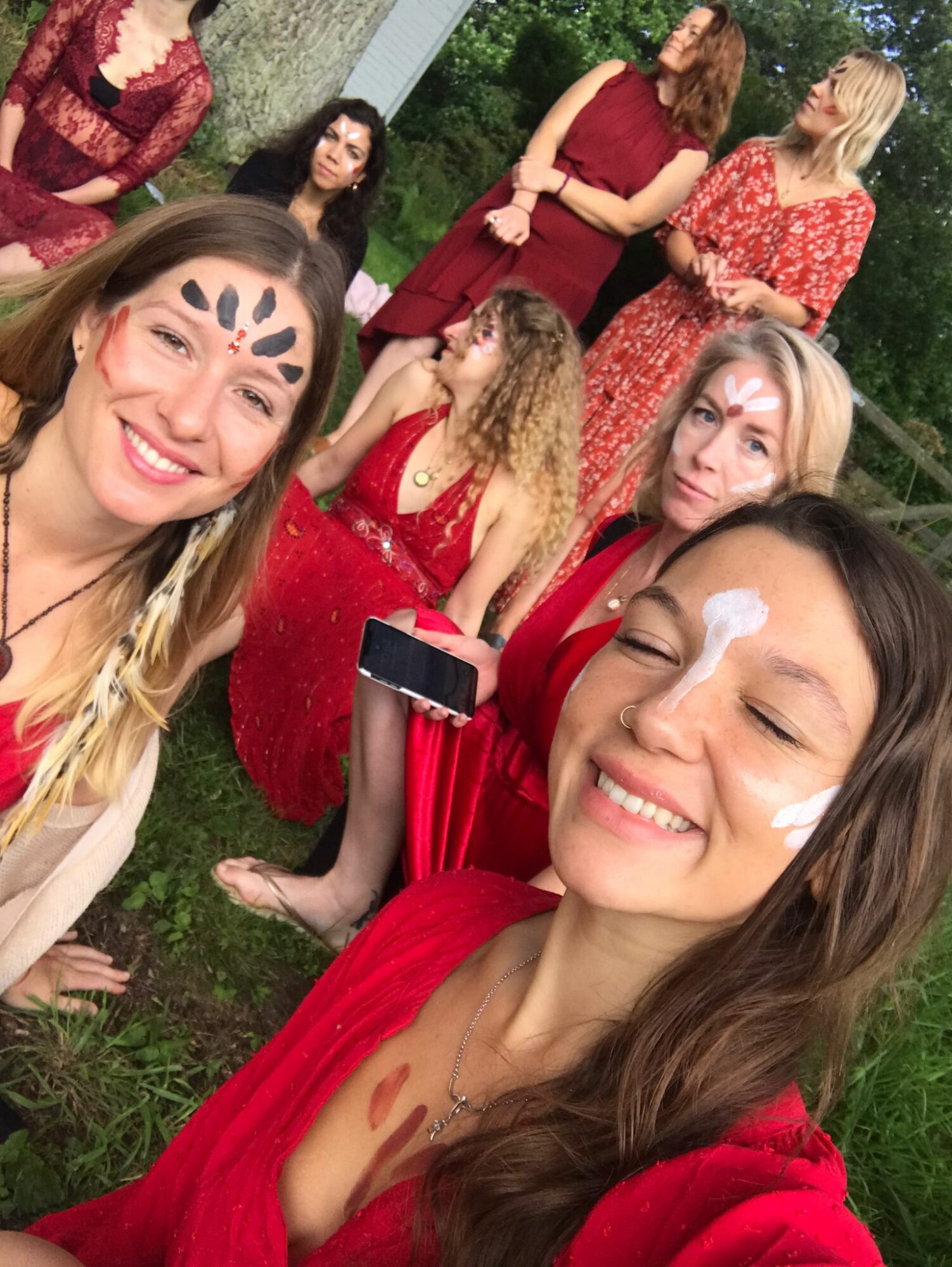 Group of women outdoors, some smiling, some talking, wearing red dresses, with trees in the background.