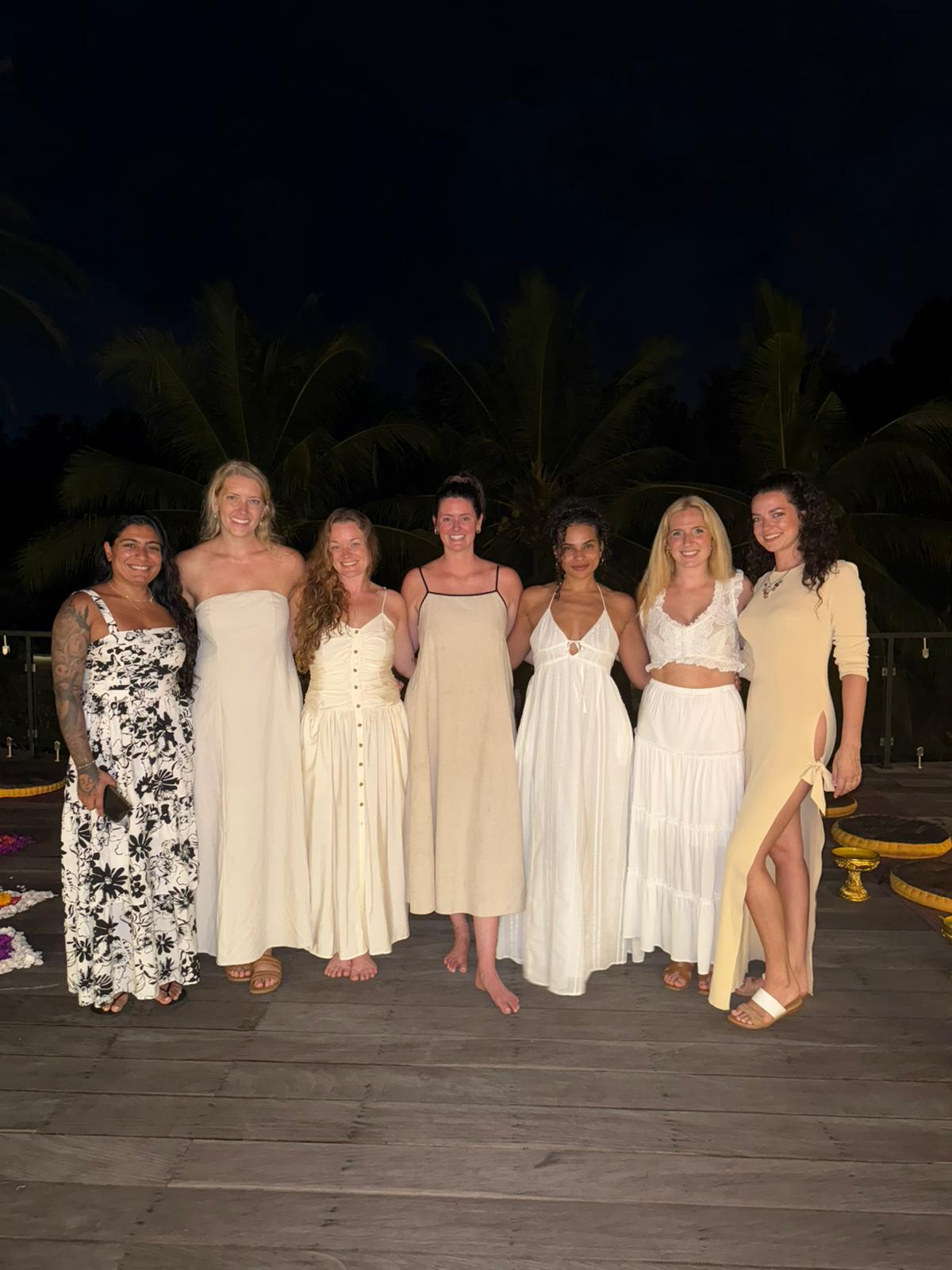 Group of seven women standing outdoors at night, wearing light-colored dresses, with palm trees in the background.
