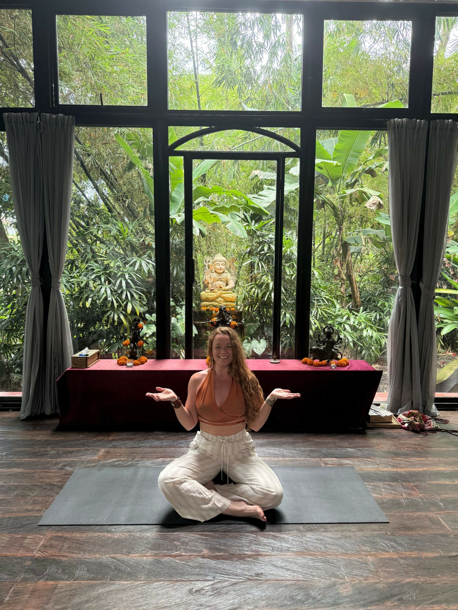 Person practicing yoga on a mat in a room with large windows and greenery outside, sitting cross-legged with arms extended.