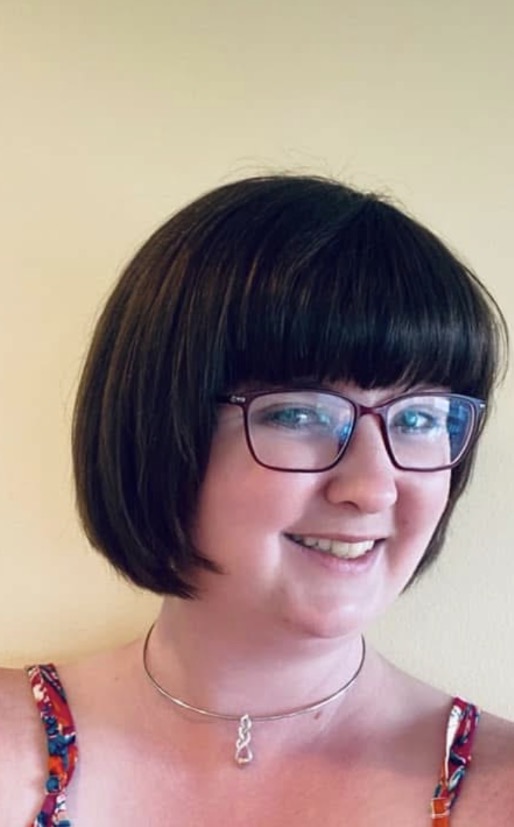 Young woman with short dark hair, glasses, smiling, wearing a colorful top and a necklace, against a plain background.