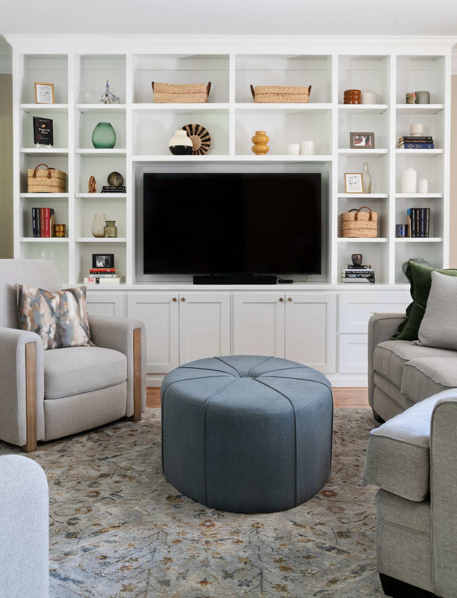 Living room with built-in white shelves, a large TV, two armchairs, a round ottoman, and decorative items.