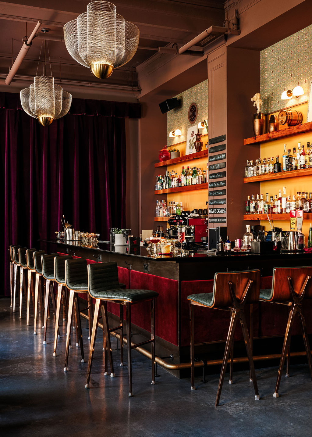 Bar with high chairs, shelves with bottles, and hanging chandeliers in a dimly lit setting.