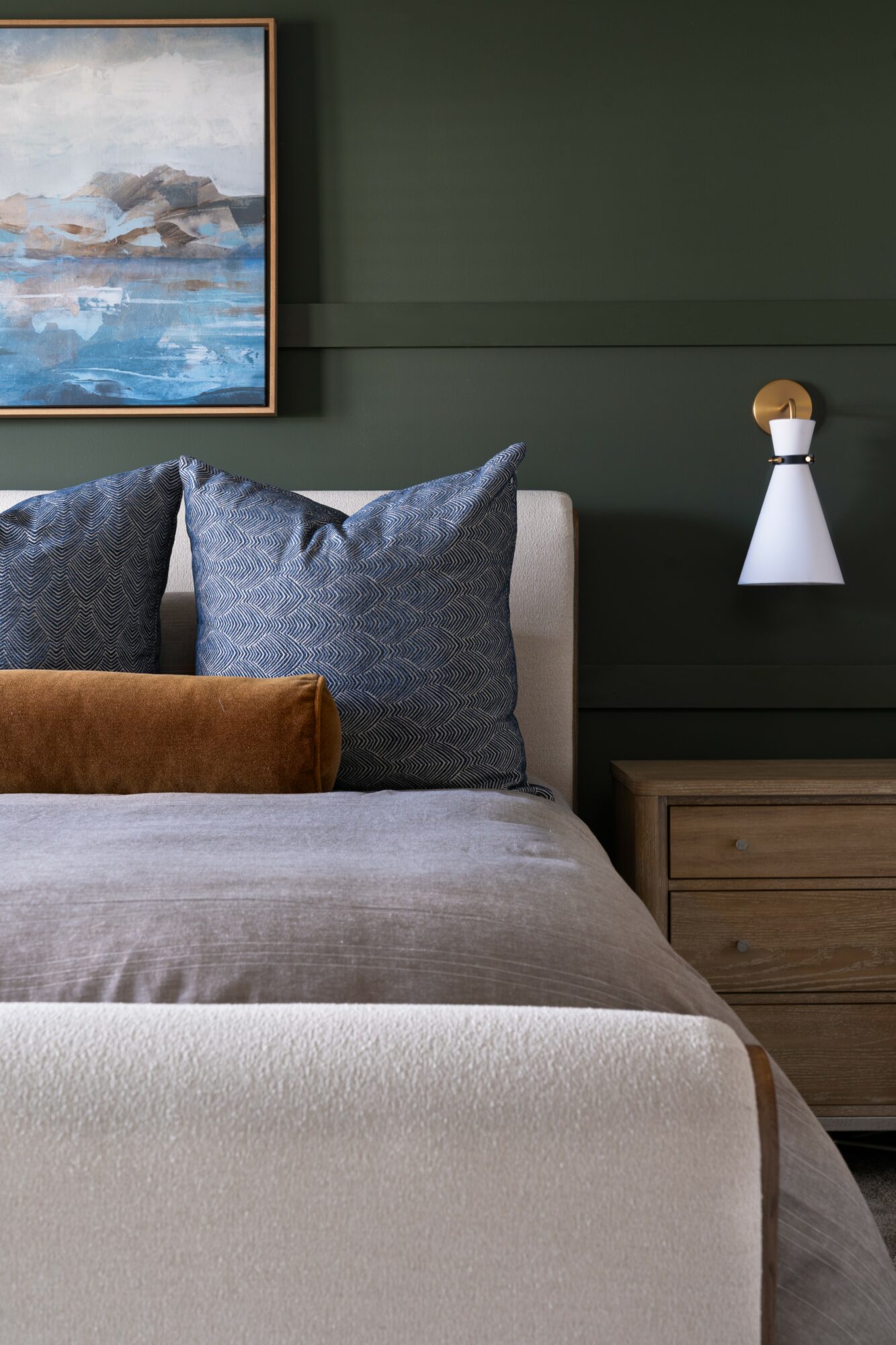 Part of a bedroom with a bed, pillows, a wooden nightstand, and a wall-mounted lamp against a green wall.