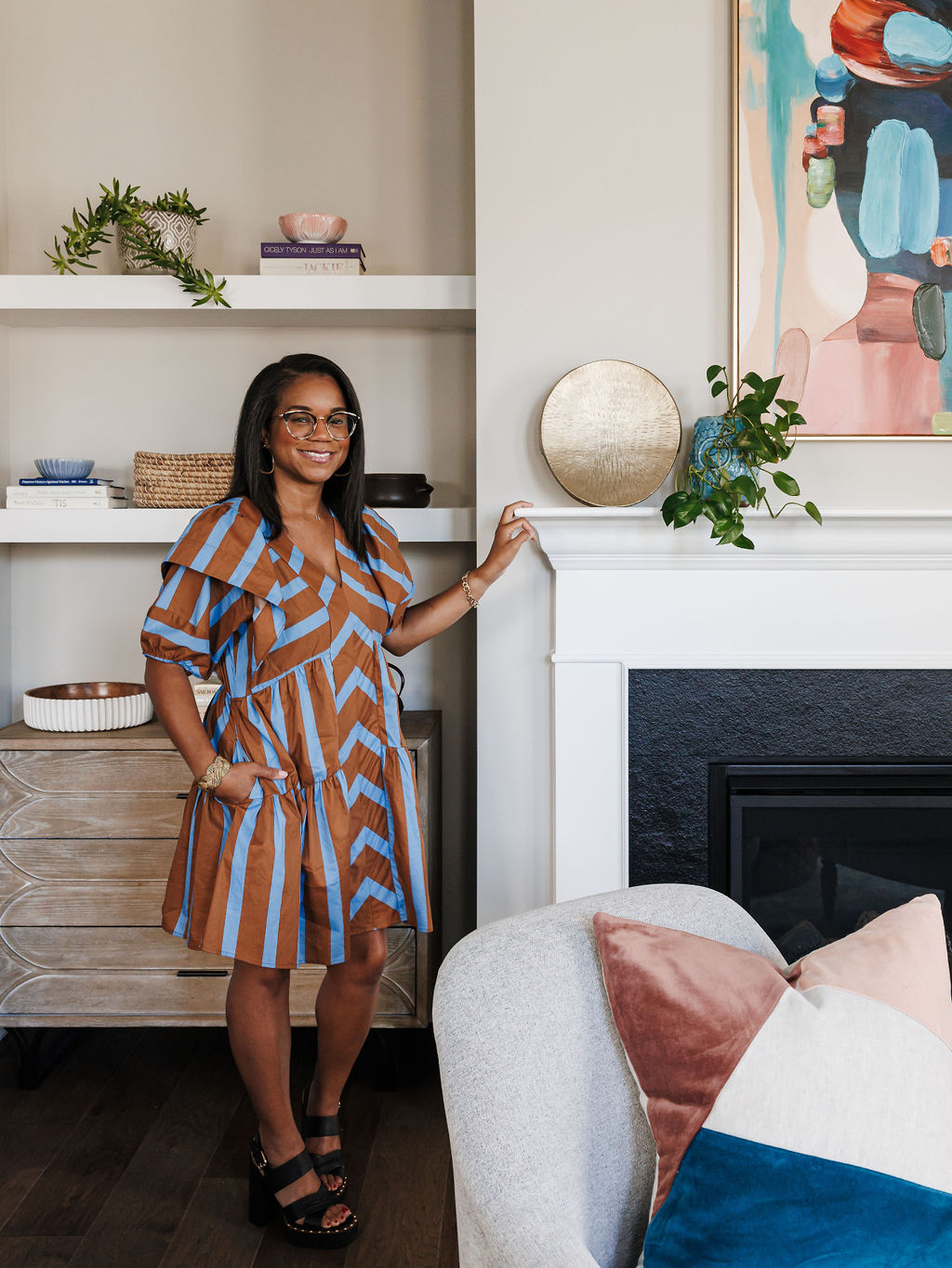 Woman with glasses in a striped dress standing near a fireplace with decorative items and artwork above.
