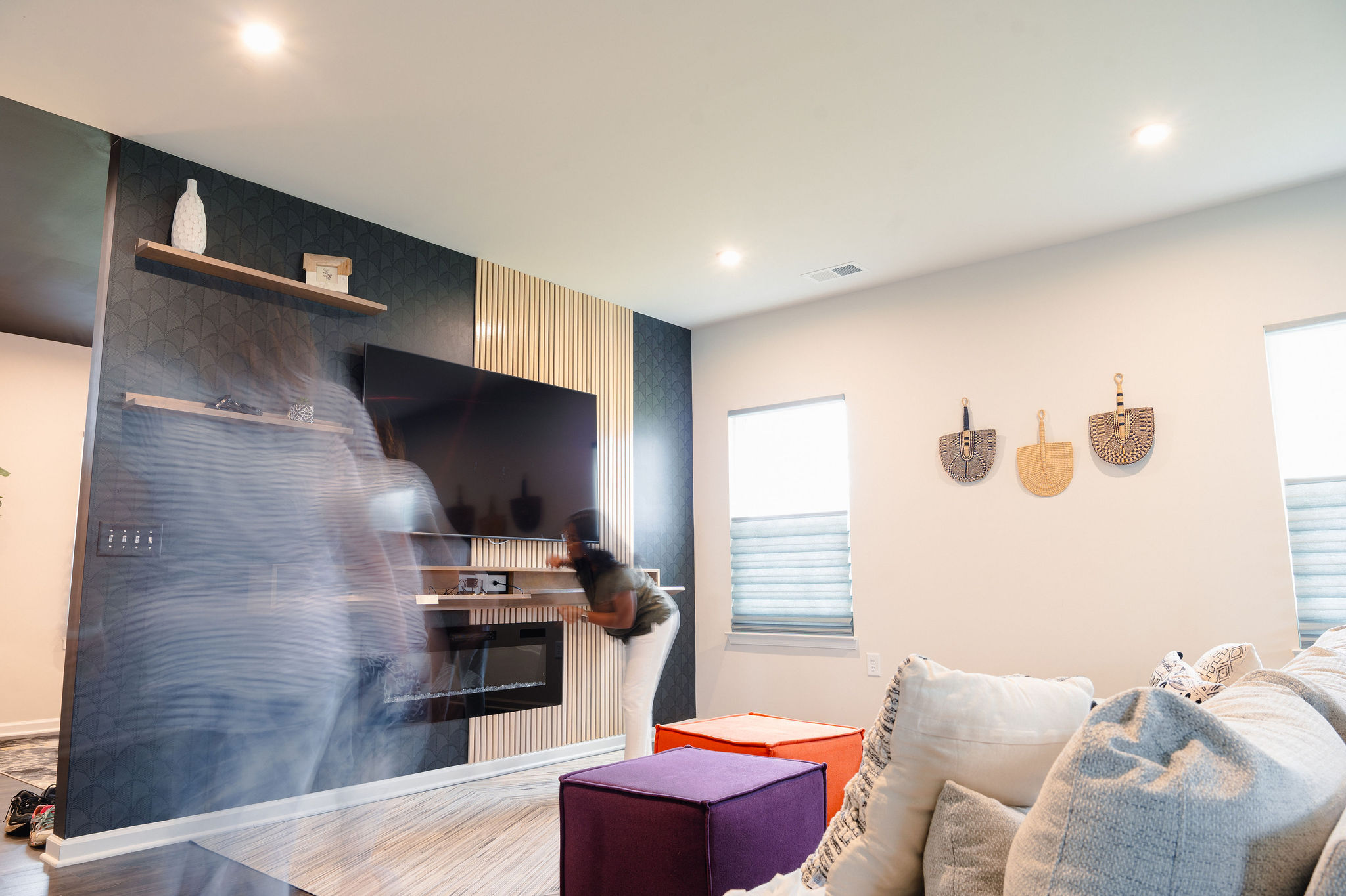 Living room with a large TV, a person adjusting the TV, and a purple ottoman, with wall decorations and pillows.