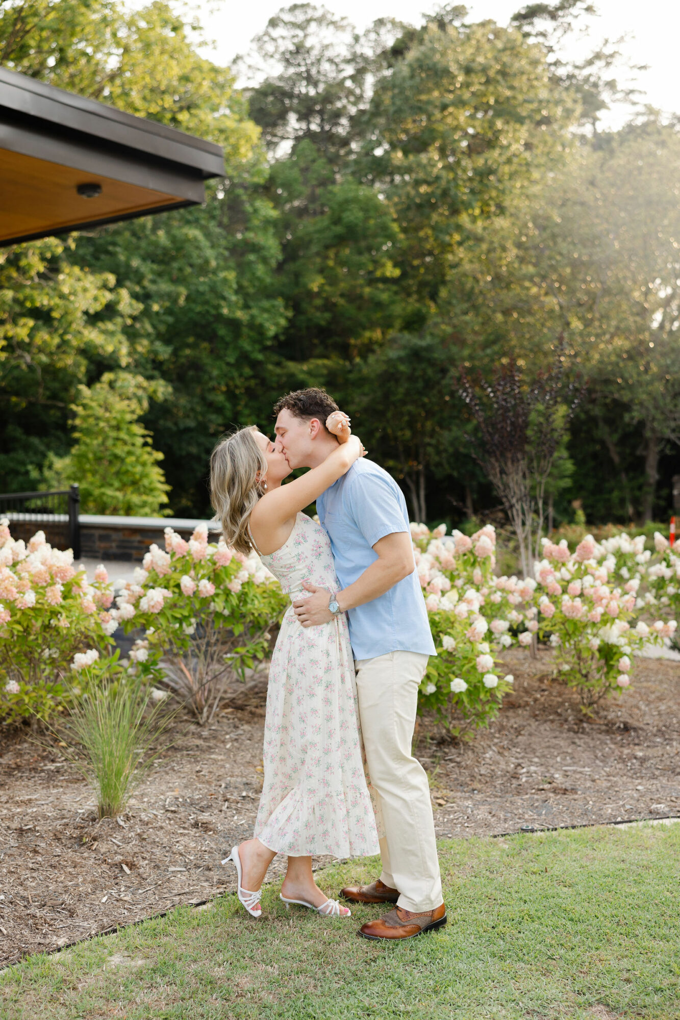 Couple kissing outdoors near flower bushes with trees in background, sunlight shining through, woman in white dress, man in light blue shirt.