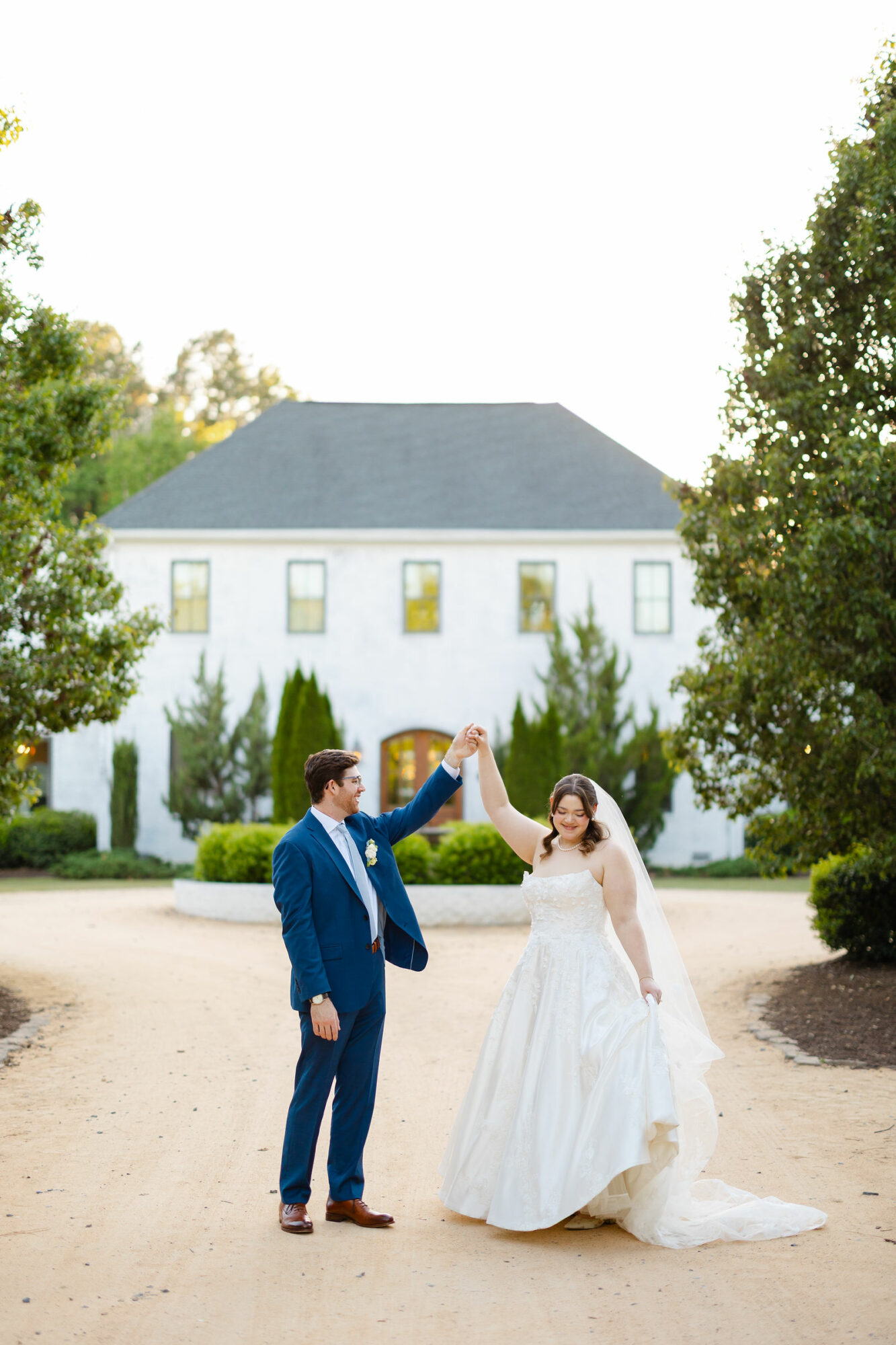 A couple in wedding attire dancing outdoors in front of a white building with trees on both sides.
