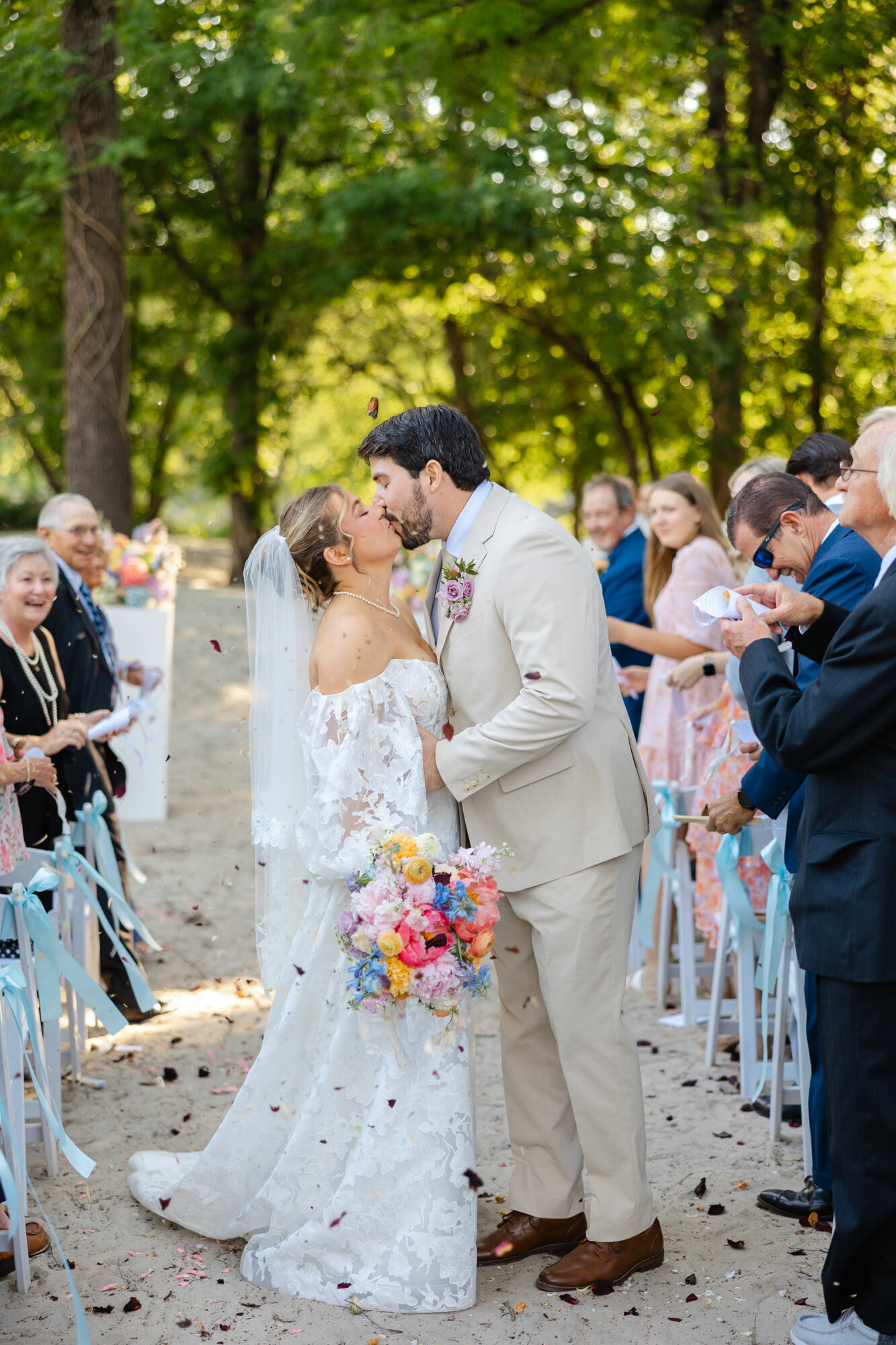 Bride and groom kiss outdoors during wedding ceremony, surrounded by guests, trees, and sunlight, bride holding colorful bouquet.
