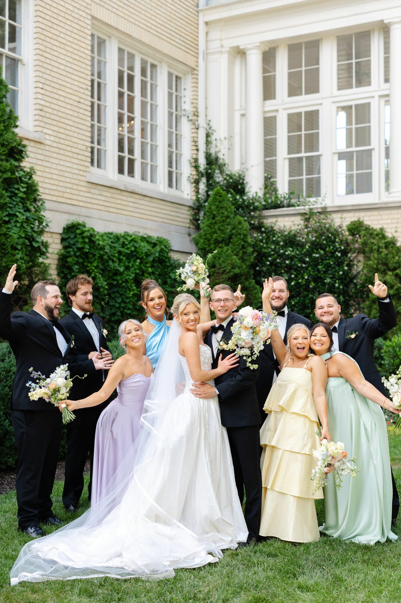 Group of people in formal attire celebrating outdoors in front of a large house with white windows, holding flowers.