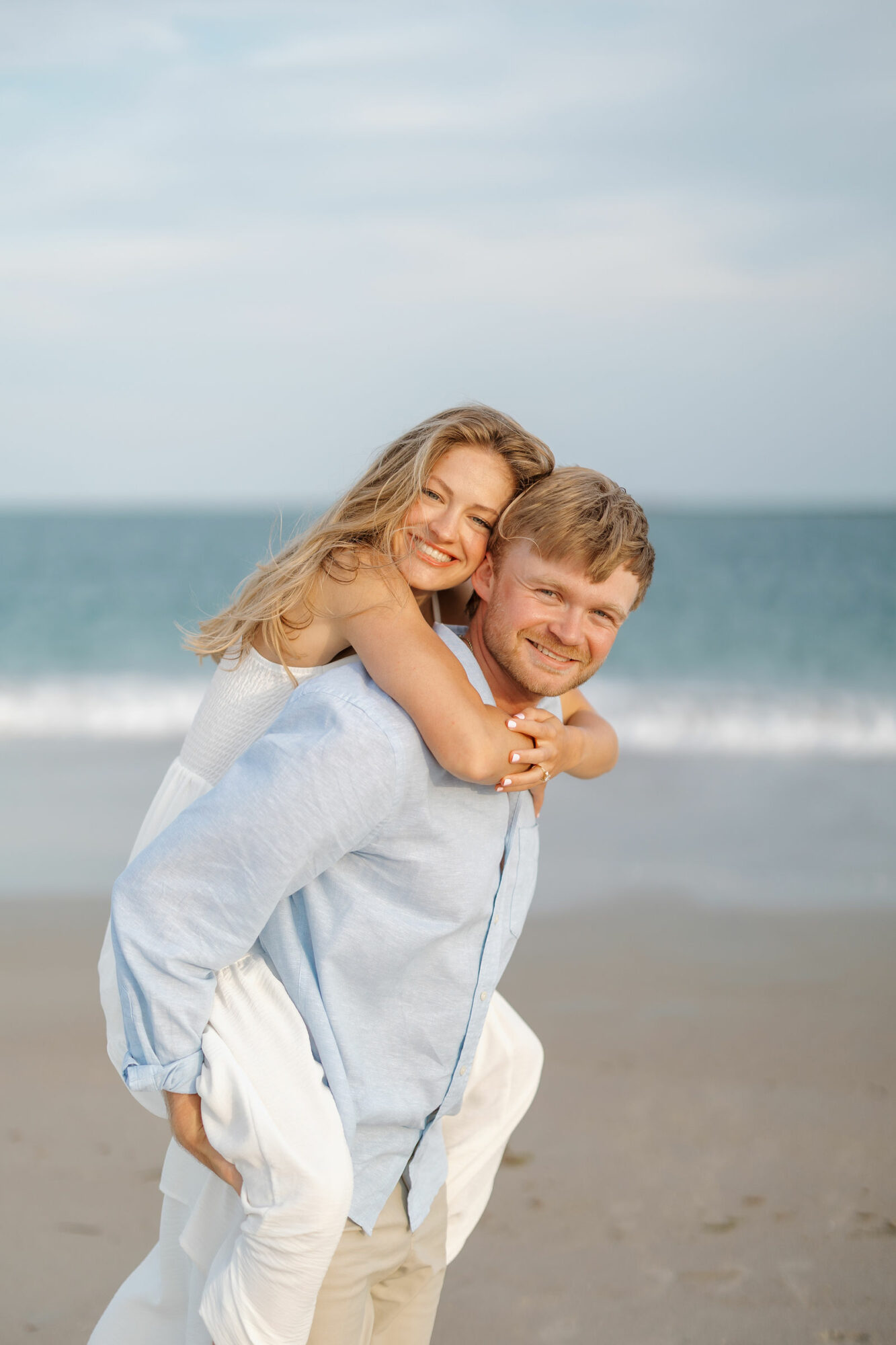 A girl and boy smiling at the beach, girl hugging boy from behind, ocean and sky in background.