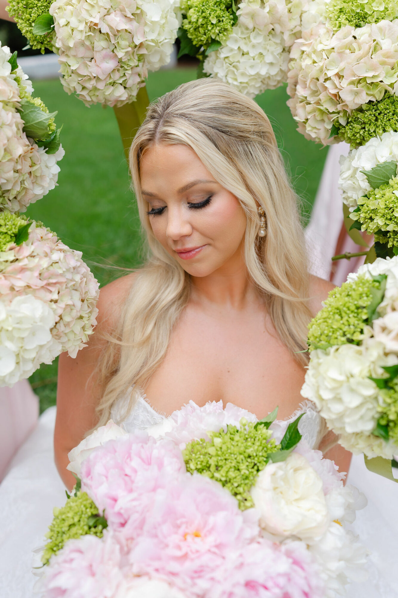 Young woman with long blonde hair in a wedding dress surrounded by white and green hydrangea flowers outdoors.