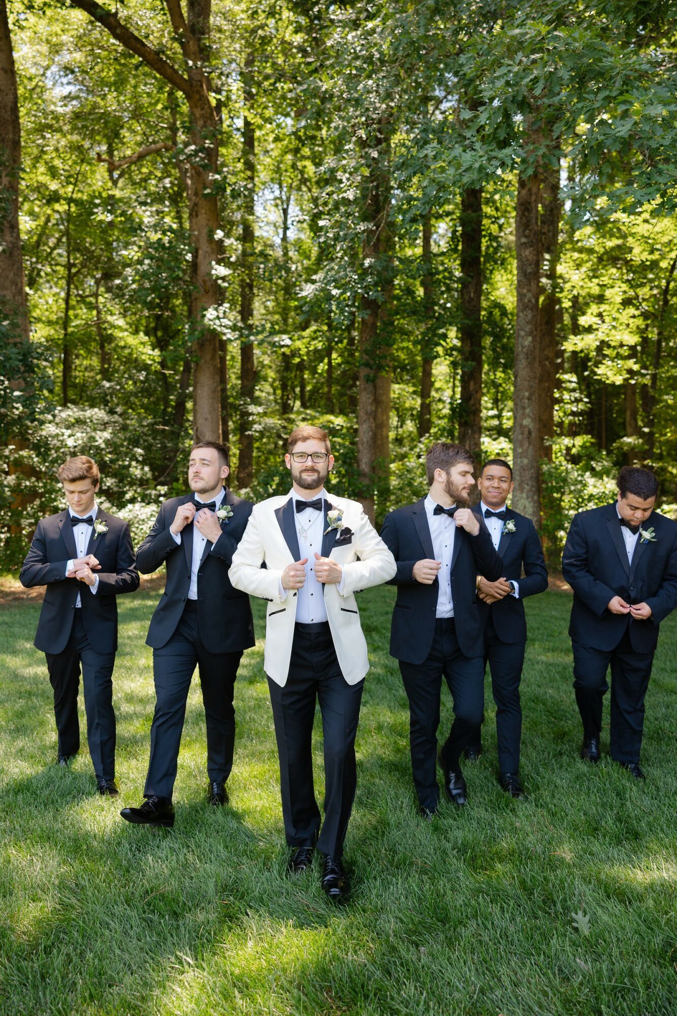 Six men in suits standing on grass in a forest, some adjusting ties, one with glasses, trees in background.