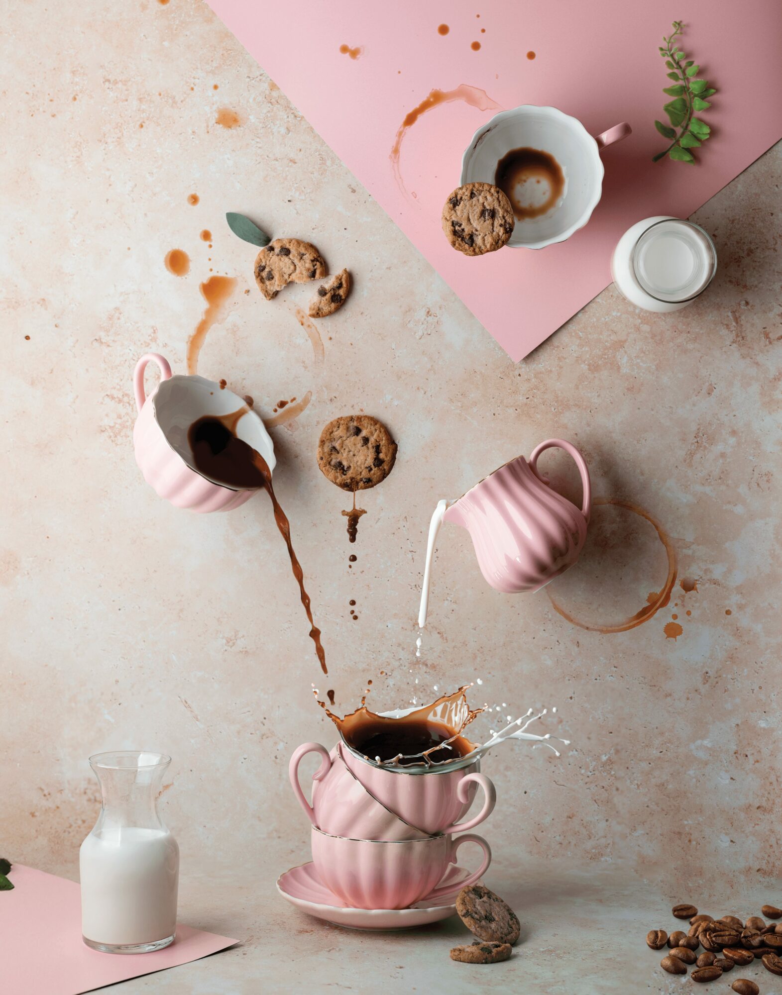 Coffee spilling from pink cups onto a saucer, with cookies, milk, and coffee beans on a textured surface and pink paper background.
