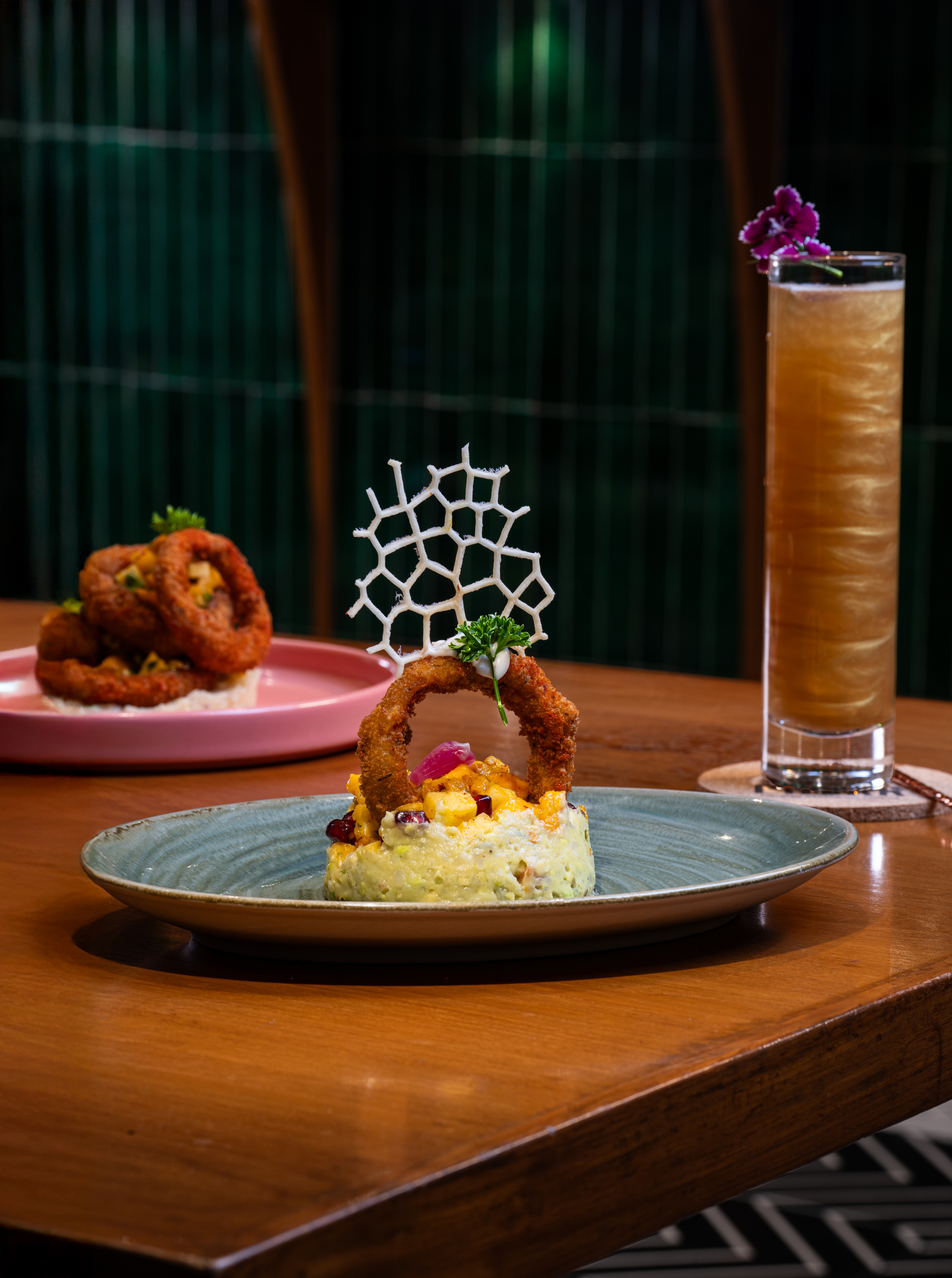 Plate with fried food, a tall drink with a purple flower garnish, and a decorative white lattice on a wooden table.